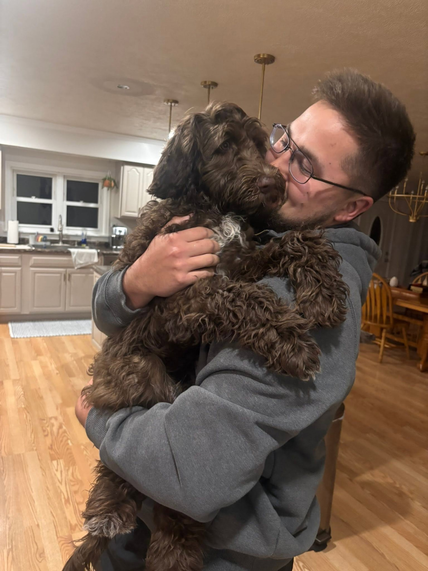 Pet dad hugging his chocolate doodle tightly in the kitchen after a long-distance homecoming